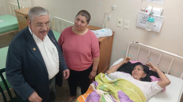 Mujer en cama con manta de colores, hombre mayor con gafas hablando, otra mujer observa seria en ambiente hospitalario.