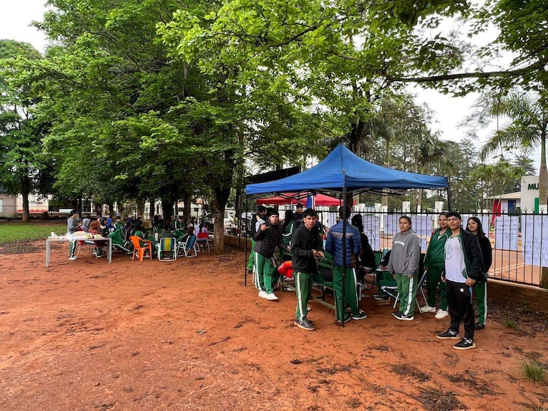 Grupo de jóvenes en ambiente exterior, vestidos mayoritariamente de verde, conversando y sentados en sillas, con carpa azul al fondo.