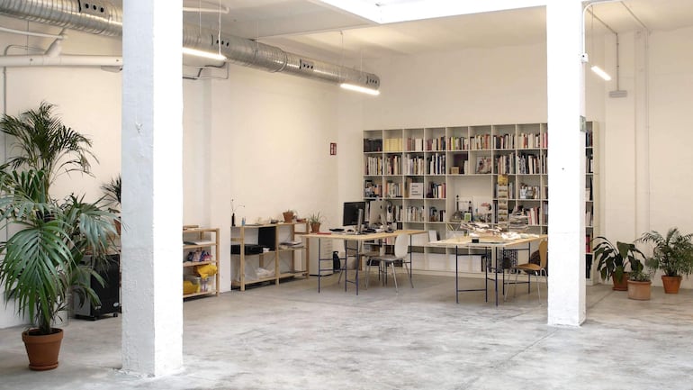Ambiente interior minimalista con mesas, estanterías llenas de libros y plantas en macetas.