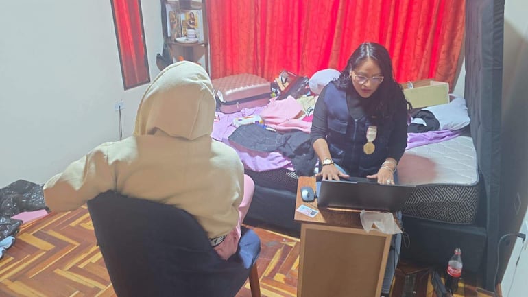 Mujer de cabello largo y oscuro con blusa negra trabaja en laptop, acompañada de una persona de espaldas en un entorno desordenado.