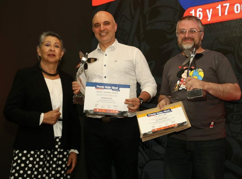Mujer canosa con blusa blanca felicita a Gustavo de Feo y otro hombre en auditorio, ambos sosteniendo trofeos.