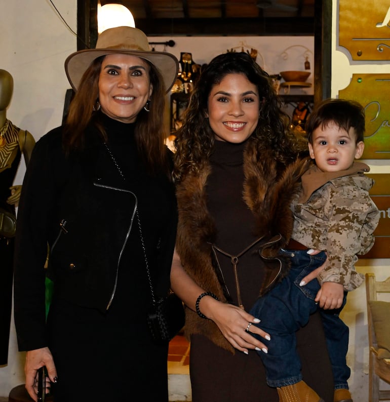 Mujer de cabello largo y oscuro con abrigo negro y sombrero claro sonríe, mientras sostiene a un niño con abrigo camuflado.