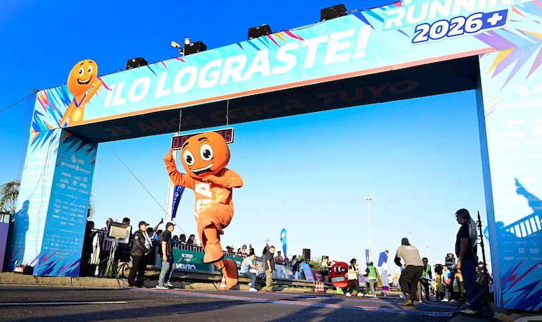 Personaje disfrazado de naranja sonríe mientras corre bajo arco de meta, con un cielo despejado y espectadores capturando el momento.