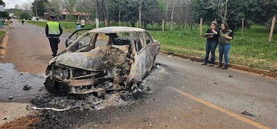 Oficial de policía con chaleco reflejante se aleja de un coche quemado, mientras dos mujeres documentan la escena con un cuaderno.