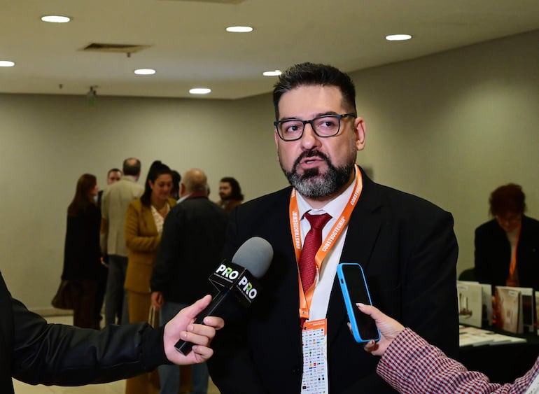 Hombre con traje negro y corbata roja, sostiene un micrófono. Asistentes atentos conversan en un salón.