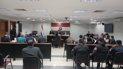 Sala de audiencias con tres miembros del tribunal y un oficial de seguridad, mientras ciudadanos observan atentos en ropa formal.