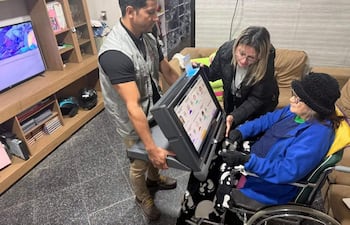 Mujer mayor sentada en silla de ruedas con abrigo azul, rodeada de dos mujeres ayudándola con un dispositivo electrónico en un espacio cálido.