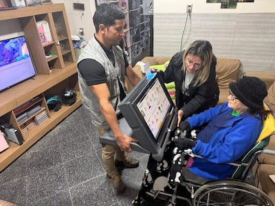 Mujer mayor sentada en silla de ruedas con abrigo azul, rodeada de dos mujeres ayudándola con un dispositivo electrónico en un espacio cálido.