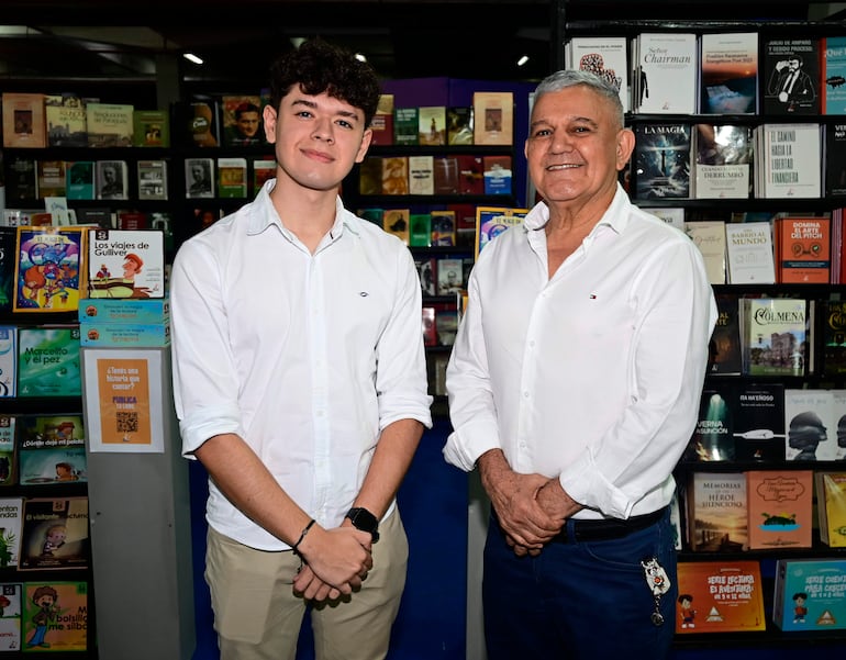 Hombres sonrientes en librería, uno con camisa blanca y pantalón claro, el otro en camisa blanca y pantalón oscuro, rodeados de libros.