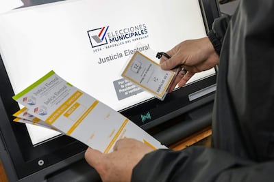 Persona con credencial electoral en mano, manipulando documentos en un entorno de capacitación electoral con pantalla informativa al fondo.