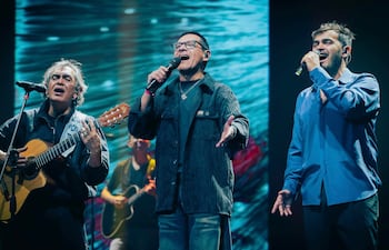 Tres hombres cantan fervorosamente en el escenario, uno sostiene una guitarra y otro levanta la mano con entusiasmo.