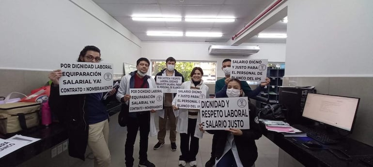 Mujer sonriente al centro sostiene cartel sobre salario digno, acompañada por seis colegas con batas blancas y mensajes similares en un ambiente claro.