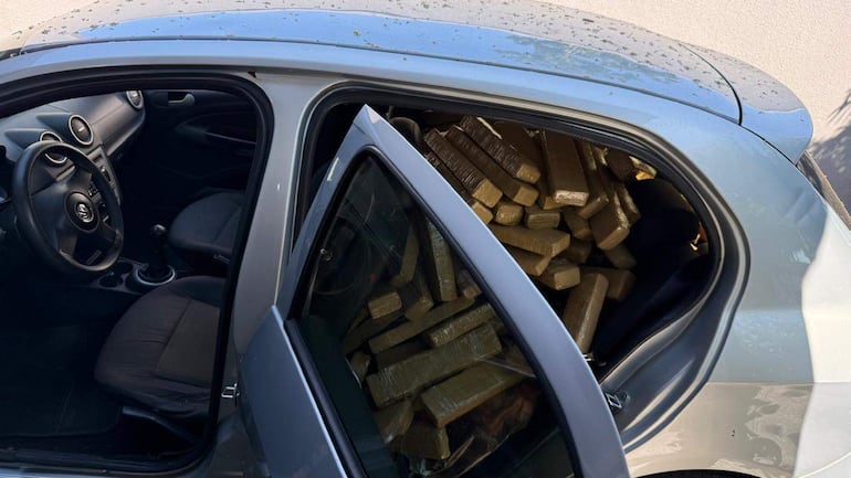 Vehículo gris con puerta abierta y paquetes marrones apilados en el asiento trasero, estacionado en un área urbana.