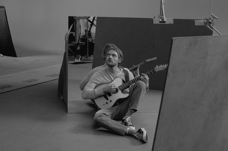 Finneas sentado en el suelo con camiseta clara y jeans, tocando la guitarra en un estudio con equipo visible.