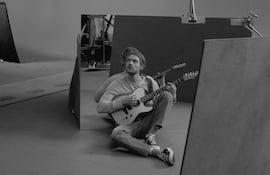 Finneas sentado en el suelo con camiseta clara y jeans, tocando la guitarra en un estudio con equipo visible.