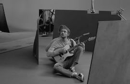 Finneas sentado en el suelo con camiseta clara y jeans, tocando la guitarra en un estudio con equipo visible.