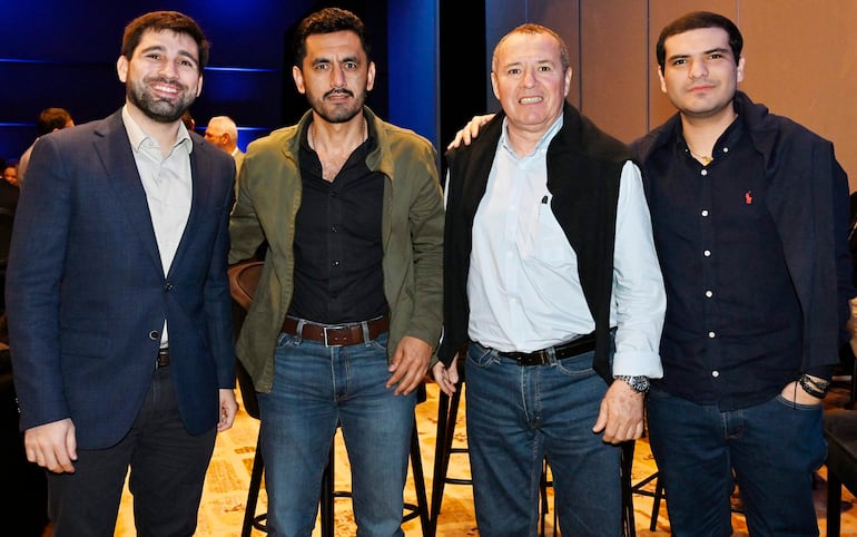 Cuatro hombres, uno sonriente con saco oscuro, otro serio en camiseta negra, y dos más con camisas claras, posan en ambiente moderno.