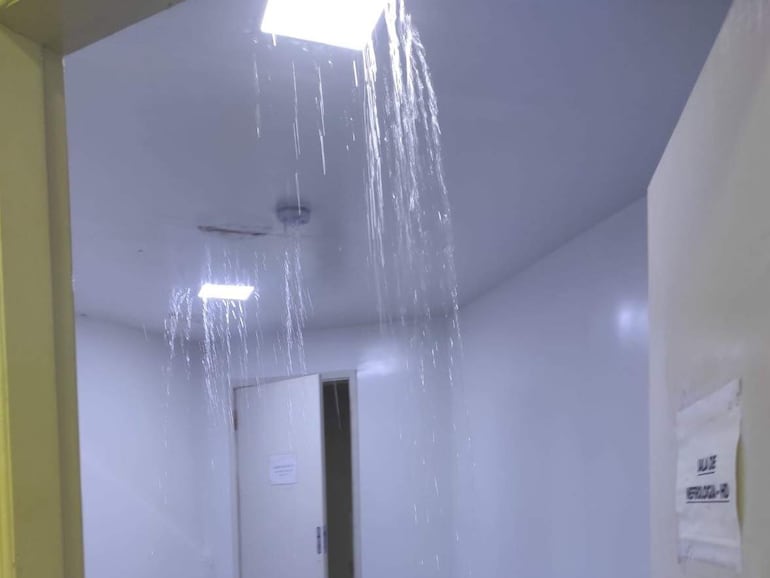 Pasillo interior del hospital con agua cayendo del techo y cartel de 'SAL DE EMERGENCIA' visible en la esquina derecha.