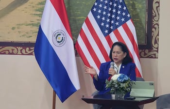 Dra. Celina Realuyo en blazer azul, hablando con micrófono, flanqueada por banderas de Paraguay y EE. UU., en ambiente de conferencia.