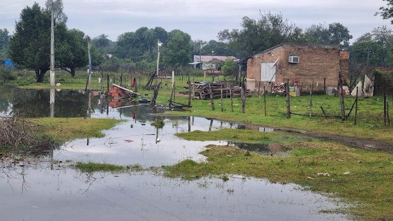 Paisaje rural inundado con vivienda de ladrillos a la derecha, vegetación densa y objetos flotantes en el agua.