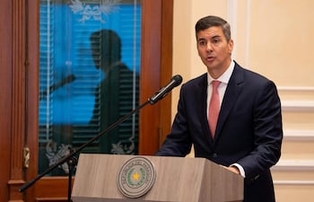 Santiago Peña Santi de pie en un podio oscuro, vestido con traje y corbata rosada, con expresión seria en ambiente formal.