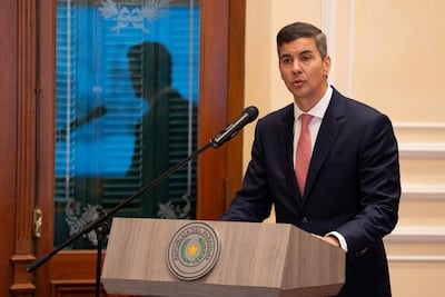 Santiago Peña Santi de pie en un podio oscuro, vestido con traje y corbata rosada, con expresión seria en ambiente formal.