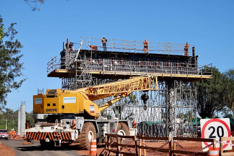 Cuatro trabajadores en cascos naranjas supervisan la construcción elevada mientras una grúa amarilla levanta materiales.
