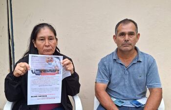 Mujer con chaqueta negra y hombre en camiseta azul, sentados con cartel de Benjamín, expresiones serias, en un ambiente público.
