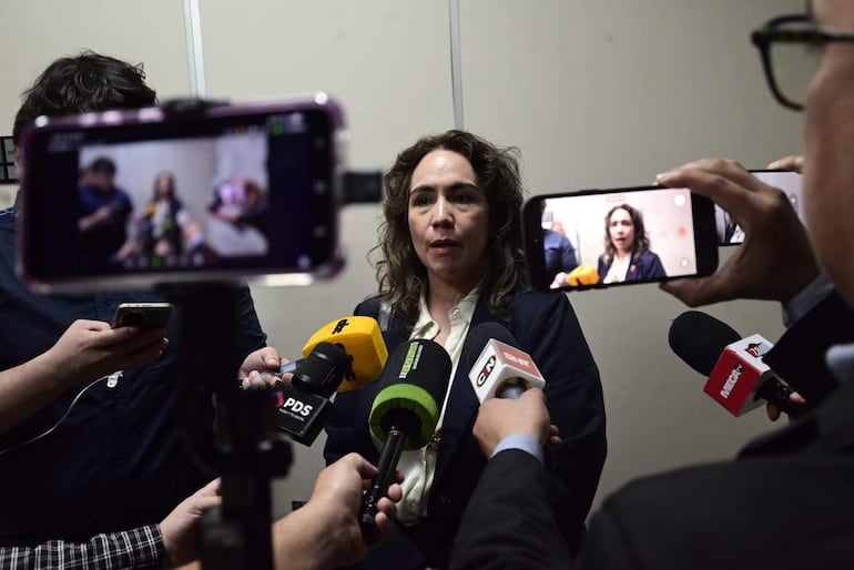 Olga Zaracho, con cabello rizado y blusa clara, rodeada de periodistas con micrófonos y teléfonos en un evento de prensa.