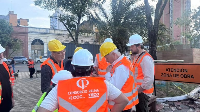 Hombres con chalecos naranjas y cascos amarillos conversan y trabajan en un ambiente de construcción, con señal de advertencia de zona de obra.