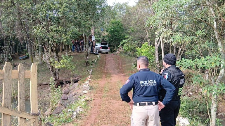 Dos policías en uniformes de investigaciones observan un grupo en el fondo, rodeados de árboles y un camino terroso.