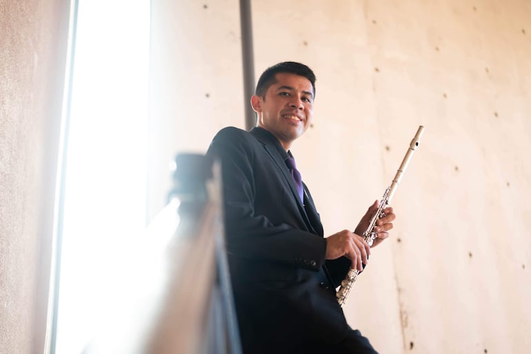 Músico en traje oscuro y corbata morada sostiene flauta, sonriendo en un entorno minimalista con paredes claras.