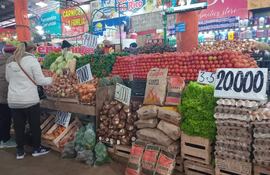 Dos mujeres de espaldas, una con chaqueta clara y mochila, observan frutas y verduras en un vibrante mercado local.