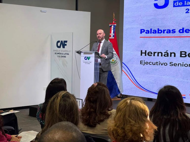Hernán Benítez en traje gris habla desde un atril con logo del CAF, con bandera de Paraguay visible y audiencia atenta.