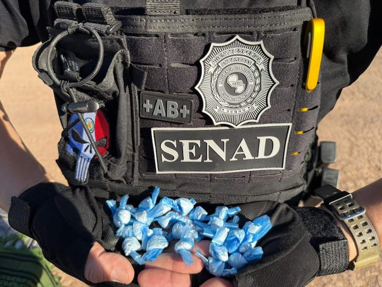 Agente antidrogas con chaleco de la SENAD sostiene paquetes azules, en fondo desértico.