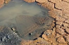 Alcantarilla rectangular sucia y desgastada, parcialmente sumergida en agua sobre pavimento de adoquines deteriorado.