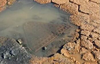 Alcantarilla rectangular sucia y desgastada, parcialmente sumergida en agua sobre pavimento de adoquines deteriorado.
