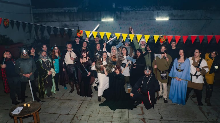 Aproximadamente 30 personas con trajes medievales levantan espadas y copas, destacando una mujer en vestido negro.