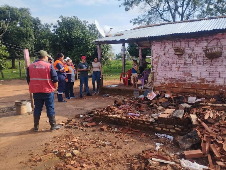 Hombre de chaleco rojo habla con grupo de personas, rodeados de árboles y escombros en ambiente rural.