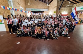 Grupo de niños y adultos con camisetas rojas y blancas, sonriendo y levantando carteles en un salón decorado.