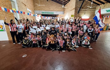 Grupo de niños y adultos con camisetas rojas y blancas, sonriendo y levantando carteles en un salón decorado.