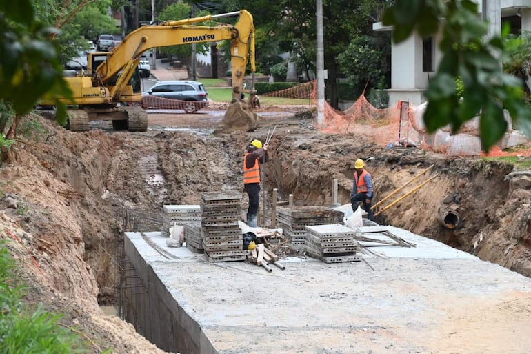 Dos trabajadores en casco amarillo y chaleco naranja en obra, mientras una excavadora Komatsu trabaja en el fondo.