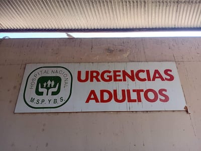 Cartel de urgencias en el Hospital Nacional, con letra roja sobre fondo blanco, parcialmente desgastado, reflejando abandono.