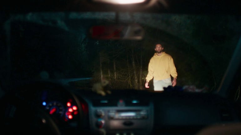 Hombre con sudadera amarilla y pantalones claros, de pie frente a un auto iluminado en un entorno oscuro y boscosa.