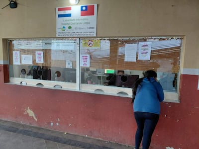 Mujer de pie con chaqueta azul apoyada en ventanal, esperando atención en el hospital, ambiente con computadoras y carteles informativos.