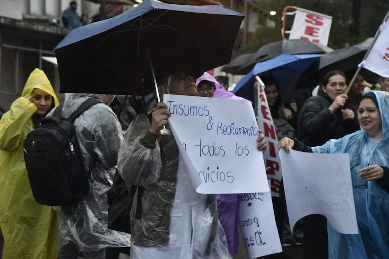 Manifestantes bajo la lluvia, algunos con impermeables y paraguas, sosteniendo un cartel que dice 'Insumos & Medicamentos para todos los servicios'.