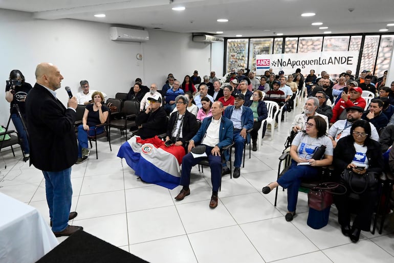 Hombre con barba frente a micrófono en sala con público, decorada con el cartel 'AND' y hashtag '#NoApaguenLaANDE'.