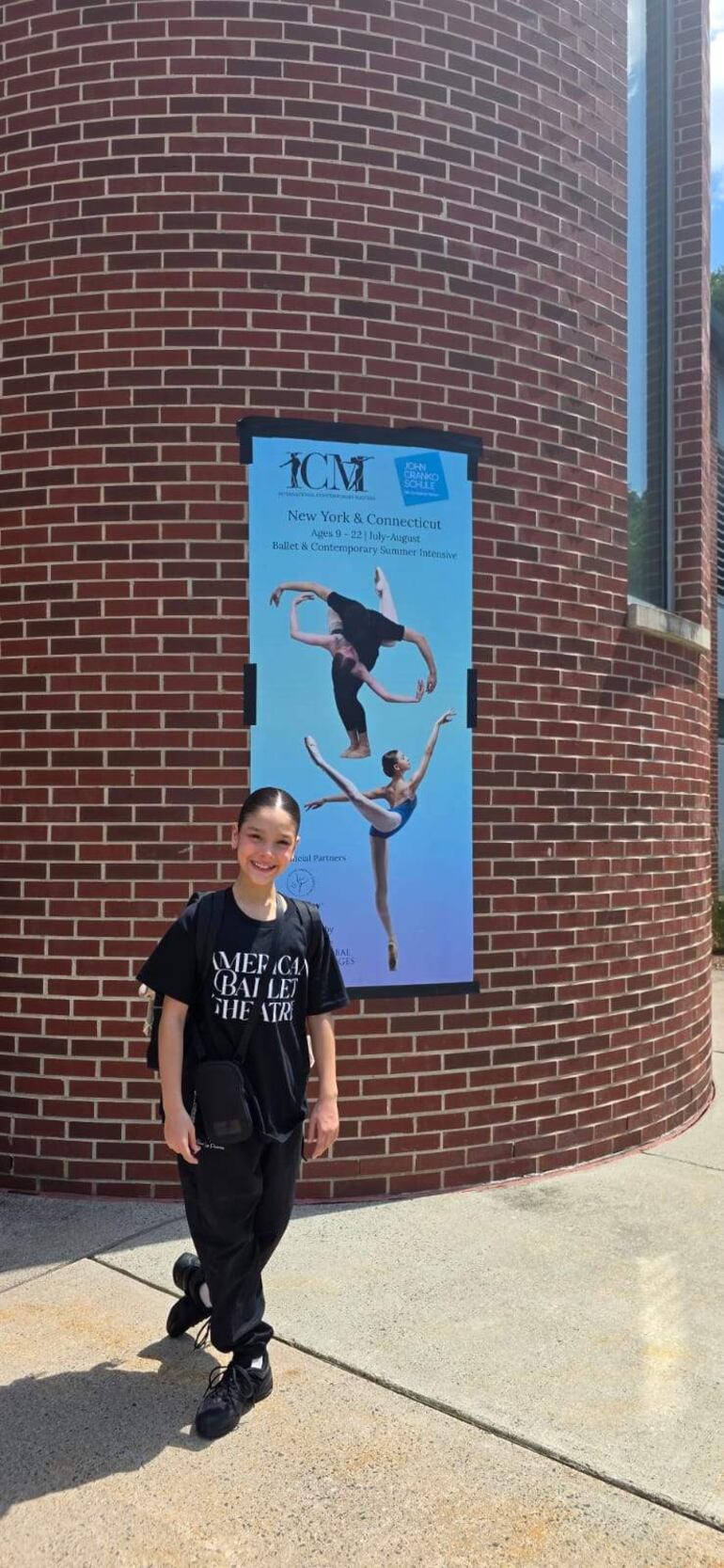 Joven con camiseta negra de American Ballet Theatre y pantalones negros, sonríe frente a un cartel de taller de danza.