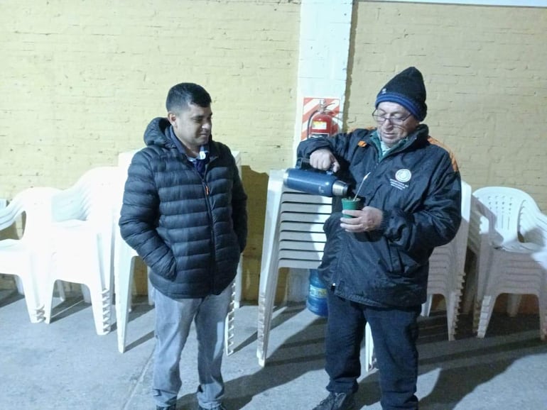 Dos hombres, uno con abrigo negro y gorro, sirven una infusión en un ambiente acogedor con sillas plásticas apiladas.