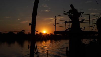 Barcos en la orilla del río al atardecer, con luz dorada reflejándose en el agua y un cielo colorido.